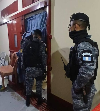 Dos hombres con uniformes militares de camuflaje y chalecos. Uno de espaldas con mochila, el otro de perfil con gorra, gafas y radio. Puerta roja y cortina