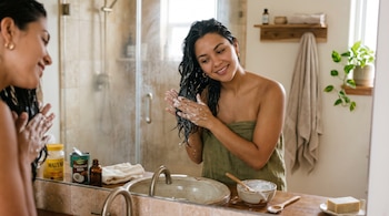 Una mujer envuelta en una toalla aplica una crema blanca en su cabello frente al espejo, junto a un tarro de maicena y una lata de leche de coco.