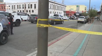 Calle urbana acordonada con cinta amarilla policial. Hay un poste de luz, dos vehículos policiales, dos oficiales y varios automóviles estacionados frente a edificios