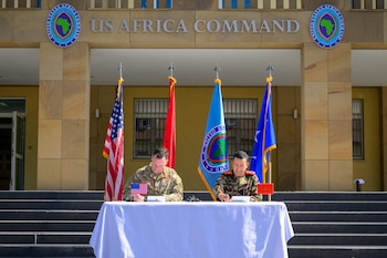 Dos militares, uno estadounidense y otro marroquí, firman documentos en una mesa con banderas de Estados Unidos, Marruecos y AFRICOM. Edificio con logo US Africa Command