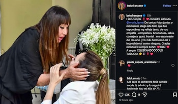 Dos mujeres, una con cabello castaño y flequillo abraza a la otra, que luce cabello castaño recogido, cerca de un florero con flores blancas