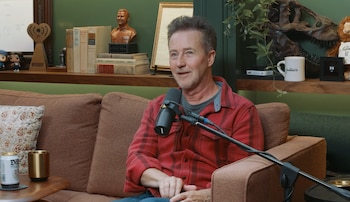 Edward Norton con camisa roja a cuadros sentado en un sofá marrón. Un micrófono lo enfoca. Detrás, estantería con libros, busto, trofeo, calavera de dinosaurio, taza, plantas. Lata y vaso en mesa