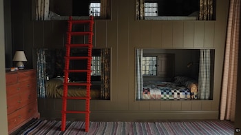 Vista de un dormitorio con paredes paneladas marrones, cuatro camas empotradas con cortinas variadas, una llamativa escalera roja central y una cómoda de madera