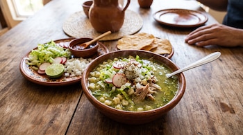 Tazón de barro con pozole verde, acompañado de tostadas, lechuga, rábanos, cebolla y limón sobre una mesa de madera.