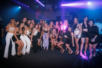 Un grupo grande de mujeres vestidas elegantemente posan sonrientes en una sala oscura iluminada con focos morados y azules