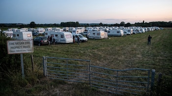 Un campo de hierba con muchas caravanas y coches, algunas personas, una señal con texto y una valla metálica. Al fondo se ven árboles y el cielo.