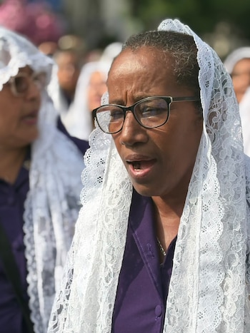 Una mujer con anteojos, hábito morado y mantilla blanca con la boca abierta, acompañada de personas con atuendos similares
