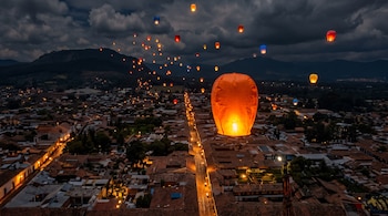 Vista aérea nocturna de un pueblo colonial con calles iluminadas y tejados, bajo un cielo nublado, con decenas de farolillos de papel de Cantoya flotando.