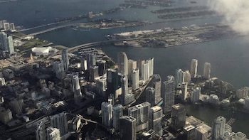 Vista aérea de rascacielos altos en una ciudad costera con puentes que cruzan una bahía, islas verdes y edificios en la distancia bajo un cielo nublado