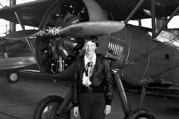 Fotografía en blanco y negro de Amelia Earhart, aviadora, de pie frente a un avión de hélice. Ella viste chaqueta de cuero y pañuelo