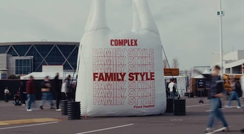 Enorme bolsa inflable blanca con texto rojo "Complex Family Style Food Festival" en un estacionamiento con personas borrosas y un edificio moderno de fondo