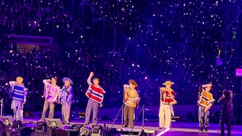 Siete integrantes de un grupo musical con ponchos y sombreros en un escenario frente a una tribuna con luces violetas