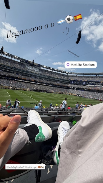 Vista parcial del MetLife Stadium con campo de césped verde, gradas con público, cielo azul, y piernas de un espectador con zapatillas blancas