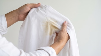 Manos sostienen una camisa de vestir blanca con manchas amarillas visibles en las dos zonas de las axilas.
