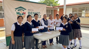 Diez estudiantes mexicanos con uniforme escolar reciben tarjetas o sobres de dos mujeres adultas sonrientes, frente a una lona del programa Beca Benito Juárez.