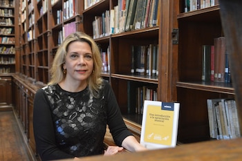 Mujer con cabello rubio, jersey negro con encaje, pendientes dorados, sentada ante estanterías de madera llenas de libros, con un libro amarillo
