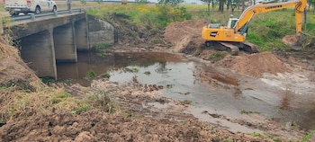 Una excavadora amarilla remueve tierra junto a un puente de concreto y un cauce con agua, mientras una camioneta blanca espera sobre el camino