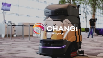 Primer plano de un robot de limpieza autónomo negro y naranja con diseño de mayordomo y una luz LED violeta, en un aeropuerto moderno con un hombre en el fondo