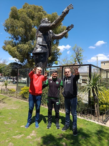 Tres hombres de pie sobre el césped posan frente a una escultura metálica de figura humana con brazos extendidos, con árboles al fondo