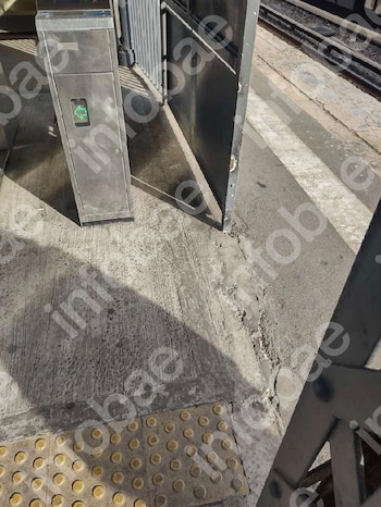 Un medidor de acceso plateado con luz verde en una plataforma de concreto. Se ve una puerta negra, baldosas táctiles amarillas y vías de tren en el fondo