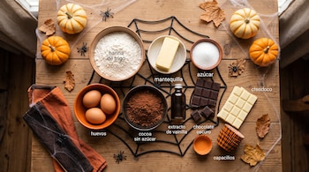 Vista cenital de recipientes con harina, huevos, cacao, azúcar, mantequilla, chocolate, calabazas y hojas secas sobre una mesa de madera.