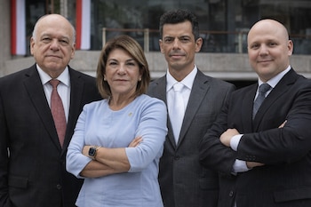 Retrato de cuatro personas: tres hombres con traje y corbata, y una mujer con blusa azul y brazos cruzados. Fondo de un edificio con ventanas.