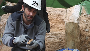 Un investigador con un casco y ropa de trabajo observa un fósil que sujeta en el mano, dentro de un yacimiento arqueológico