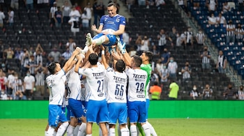Un grupo de jugadores de fútbol, vestidos con camisetas azules y blancas, levanta a un compañero en el aire en un campo de césped con gradas y público.