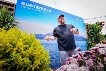 Hombre barbudo con gorra, camiseta y bermudas frente a un banner azul que dice 'GUATEMALA LAGO DE ATITLÁN, SOLOLÁ'. Arbustos y flores en primer plano