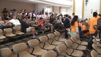 Salón de convención con una audiencia diversa de personas sentadas en sillas de patrón y algunas de pie. Algunas personas visten camisetas naranjas y rosadas