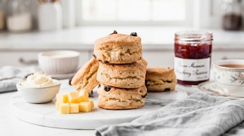 Pilas de scones con pasas, cubos de mantequilla, un tazón de crema, un frasco de mermelada y una taza de té sobre una tabla de mármol blanco.