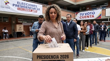 Una mujer con cabello rizado deposita su voto en una urna que dice 'PRESIDENCIA 2026' en un centro de votación en Colombia, con personas en fila y pancartas al fondo.