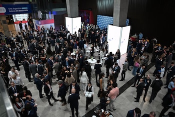 Amplio salón de eventos concurrido por decenas de personas de pie y sentadas. Hay mesas redondas, paneles luminosos verticales y personal de servicio visible