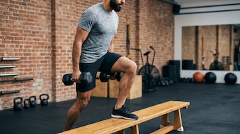 Hombre con barba y camiseta gris realiza subidas al banco con mancuernas. Se observan equipos de gimnasio, pared de ladrillos y espejo.