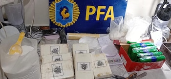 Múltiples paquetes envueltos y bolsas con polvo blanco, una balanza digital y una pala amarilla, frente a un cartel azul con el logo de la Policía Federal Argentina