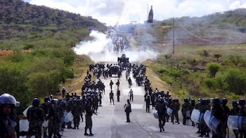 Vista aérea de una carretera con policías antidisturbios y manifestantes bajo una nube de humo blanco, rodeada de colinas verdes y un cielo nublado