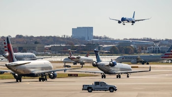 Varios aviones de pasajeros en pista de aeropuerto. Uno azul oscuro en el aire, otro blanco con cola Delta, y American. Un vehículo de servicio