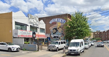 Calle urbana con edificios, un mural con "Río de la Plata" y figuras de hombres, señales de restaurante y farmacia, varios vehículos, un árbol y cielo nublado