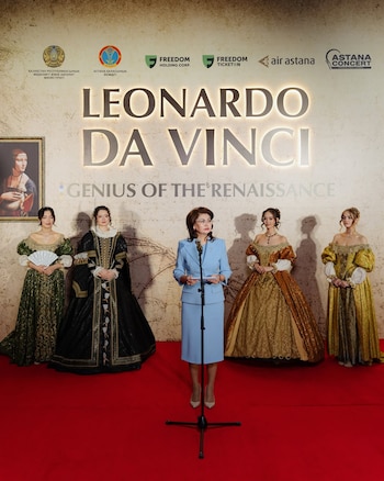 Una mujer en traje azul habla en un micrófono en una alfombra roja, junto a cuatro mujeres con trajes renacentistas, frente a un cartel 'Leonardo Da Vinci'