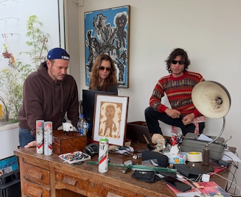Tres personas junto a un escritorio de madera repleto de latas de aerosol, un cuadro enmarcado y una máquina de escribir