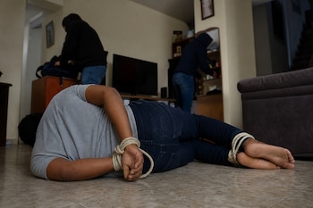 Un hombre yace en el suelo con las manos y los pies atados con cuerdas mientras dos personas con capucha buscan en los muebles al fondo.