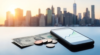 Un billete de un dólar, monedas estadounidenses y un teléfono móvil con un gráfico de línea ascendente frente a un horizonte urbano difuminado.
