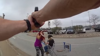 Vista desde cámara corporal de policía con arma apuntando. Mujer con cabello rojo sostiene objeto afilado junto a niño en carrito de compras en estacionamiento