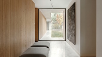Pasillo moderno con pared de madera laminada, dos bancos grises, gran ventanal central a árboles exteriores y un cuadro oscuro en pared derecha.