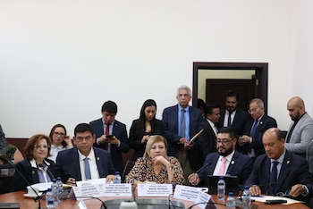 Un grupo de hombres y mujeres, muchos en trajes formales, sentados alrededor de una mesa de conferencias con micrófonos y botellas de agua