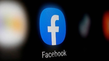 Primer plano del logo azul de Facebook con una 'f' blanca, visible en una pantalla oscura, con la palabra 'Facebook' escrita debajo y luces borrosas al fondo