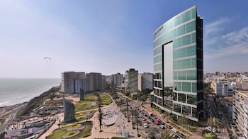 Vista aérea de la costa de Lima con el hotel JW Marriott, edificios modernos, el océano Pacífico, un parque verde, tráfico y un parapente en el cielo despejado