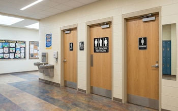 Tres puertas de madera con carteles de baños para hombres, todos los géneros y mujeres. Un bebedero doble y dispensador de gel hidroalcohólico en la pared. Un espejo refleja casilleros azules