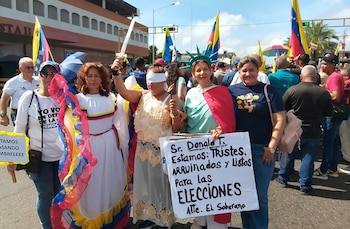 Manifestantes en una calle. Una mujer con venda en ojos y espada. Otra con corona de Estatua de la Libertad. Banderas de Venezuela y pancartas visibles