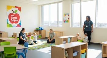 Un aula espaciosa con grandes ventanales, niños pequeños jugando, dos adultos supervisando, mobiliario infantil colorido y un gran cartel con el logo 'The Little Apple'.
